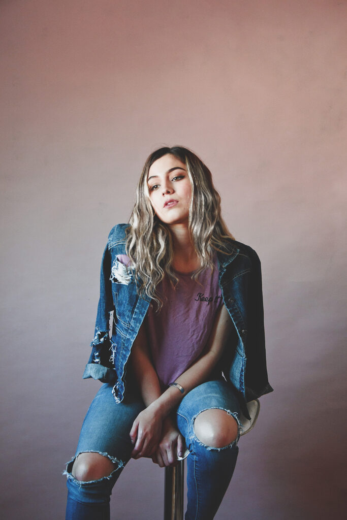 Darian Rojas a model with wavy blonde hair sits against a muted pink backdrop wearing a denim jacket with patches a purple t-shirt and distressed blue jeans.